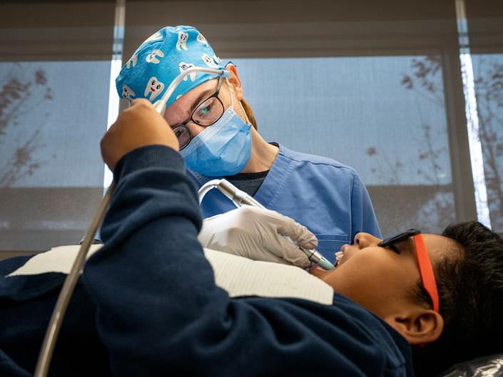 A dental student works on a child wearing protective eyewear in a dentist's chair.