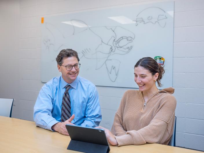 A professor sits at a table with a student as they discuss work on a laptop.