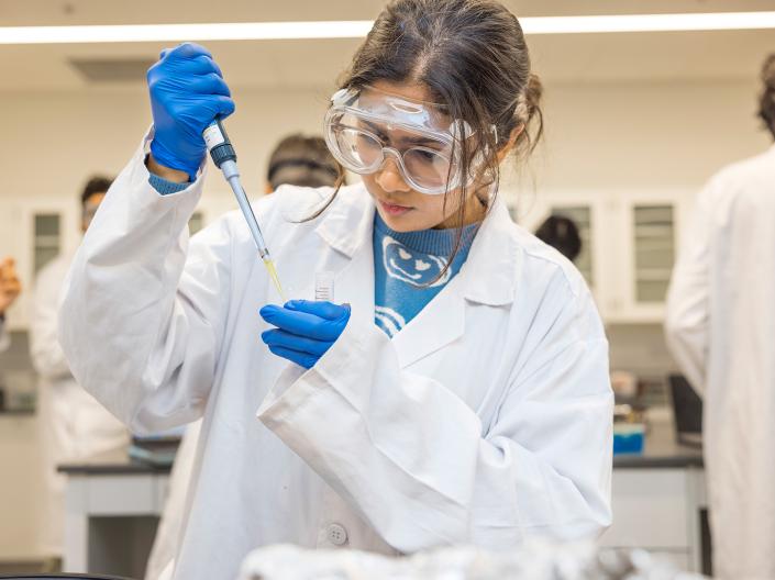 A student in a biology lab wearing PPE and adding liquid from a pipette into a test tube.