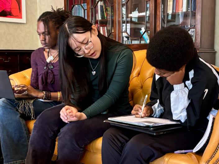 A tutor sits on a couch with two students as they review lesson work together.