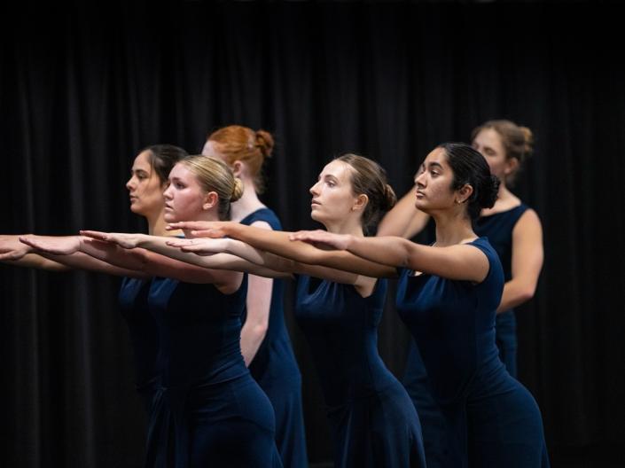 A group of dancers wearing matching dark blue dresses stand in a row with their arms extended straight forward against a dark background.