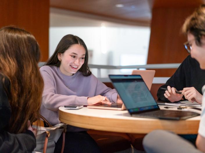 A group of students smiling and collaborating around a table with laptops in a modern indoor campus setting.