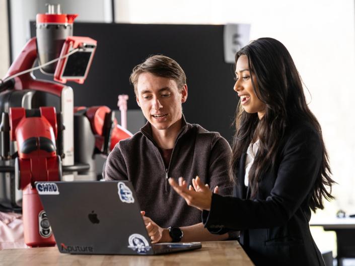 Two students talk and work on a laptop while robotics hang behind them.