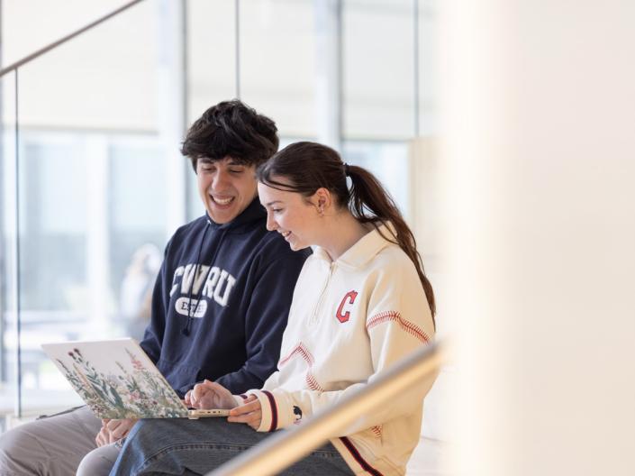 Two students sit together indoors and smile while looking at a laptop computer.
