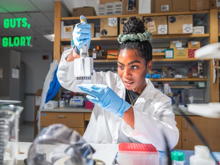 A female scientist in a white lab coat and blue gloves using a multichannel pipette in a genetics research laboratory.