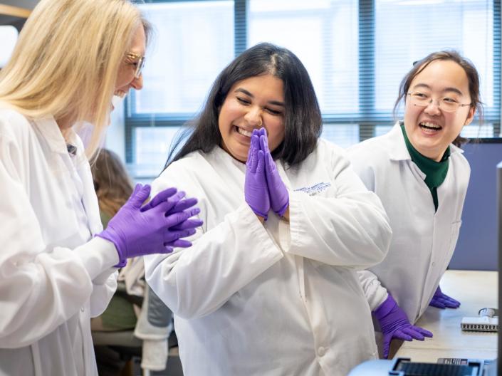 Three researchers in PPE smile and clap as they celebrate in their lab.