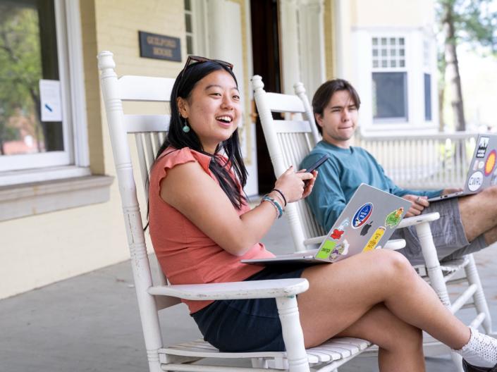 Two students sit in white rocking chairs on a building porch while using their laptops and phone.