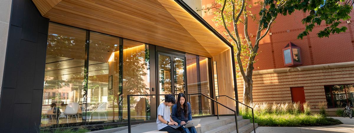 Two students sitting on a bench outside a university building with a wooden canopy, working on their laptops.