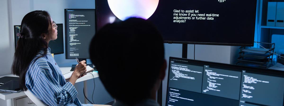  Two researchers in a dark office working on multiple monitors displaying code and an AI interface with a voice waveform.