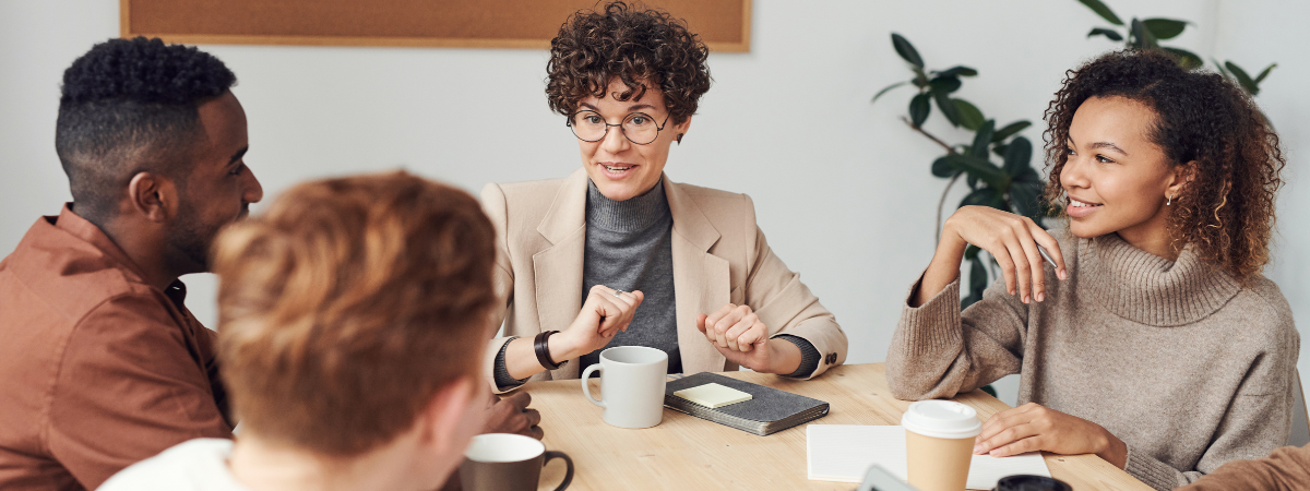 Four business professionals sit around a table talking with coffee cups and notebooks in front of them. 