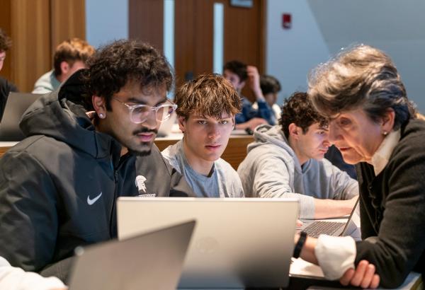 Students and a professor review work on a laptop during class.