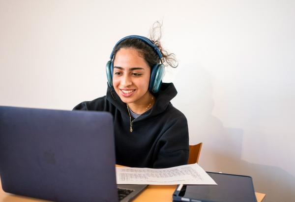 A student with headphones on smiles as she works on the laptop.
