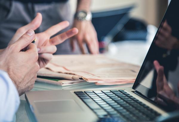 A close-up of an open laptop while two individuals talk.