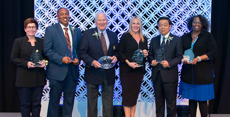 Photo of the 2018 Alumni Award Recipients, holding their awards, standing on the stage during Homecoming 2018 Weekend at the All Alumni Luncheon