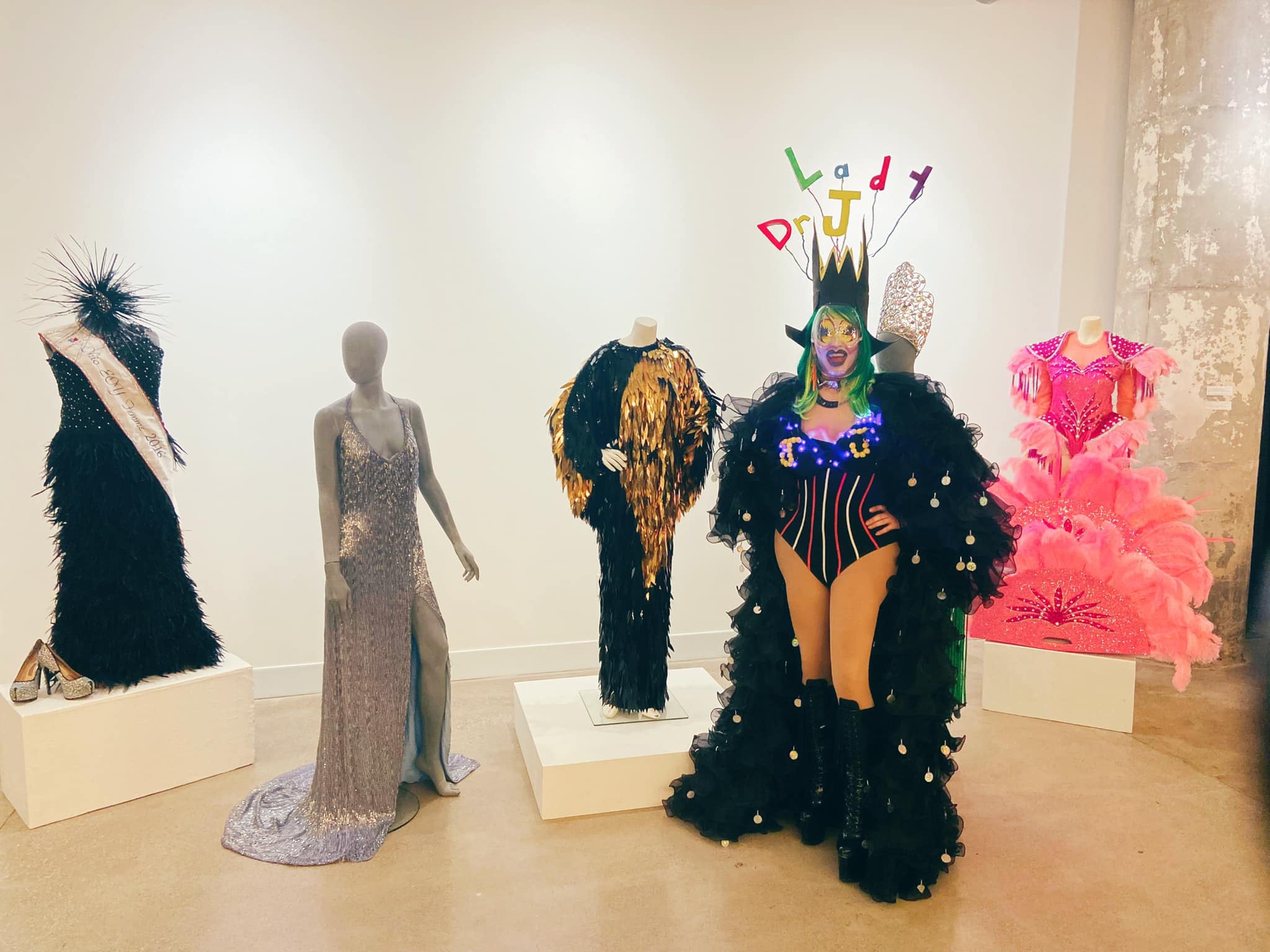 Dr. Lady J stands in full drag with her name spelled out over her head, in front of glamorous historic costumes at a drag exhibit.