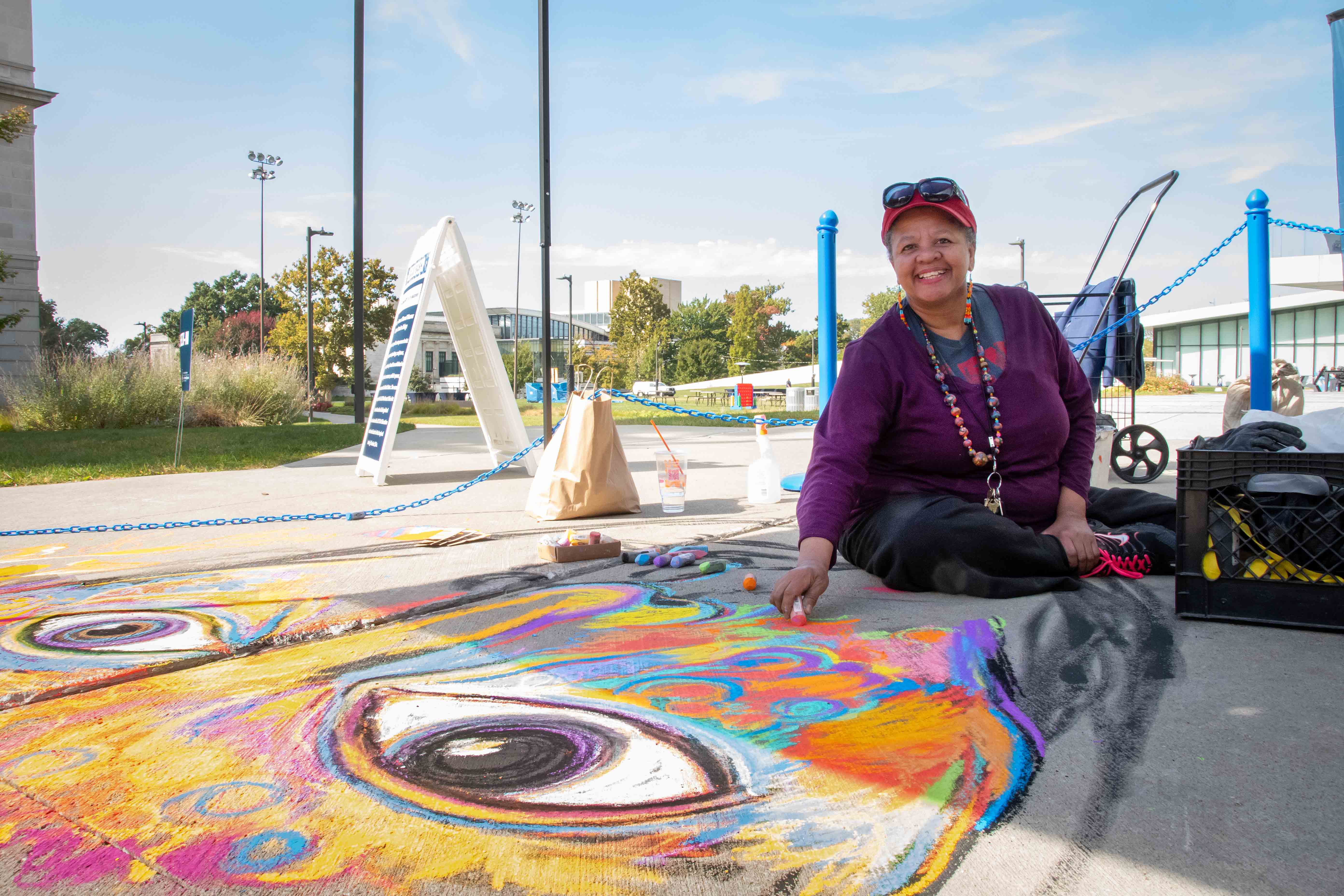 Anna sits on the ground toward the bottom of her mural with a piece of chalk in-hand, smiling at the camera. She's wearing a red hat, purple sweater and the sky behind her is blue. The portrait is a mixture of many bright colors.