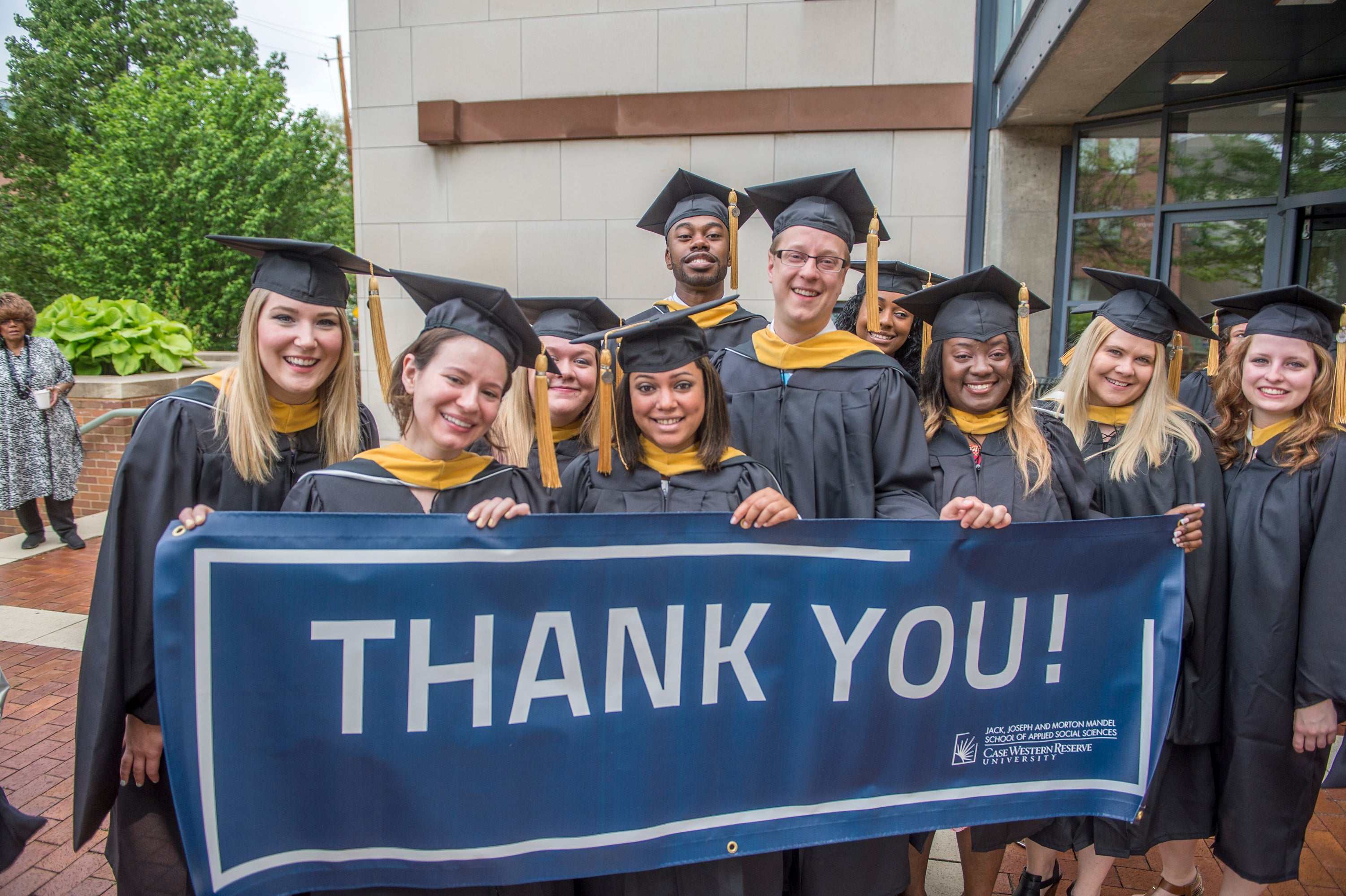Graduates from the Mandel School of Applied Social Sciences at CWRU pose in caps and gowns, smiling while hold up a blue sign that reads "Thank You!" in all caps