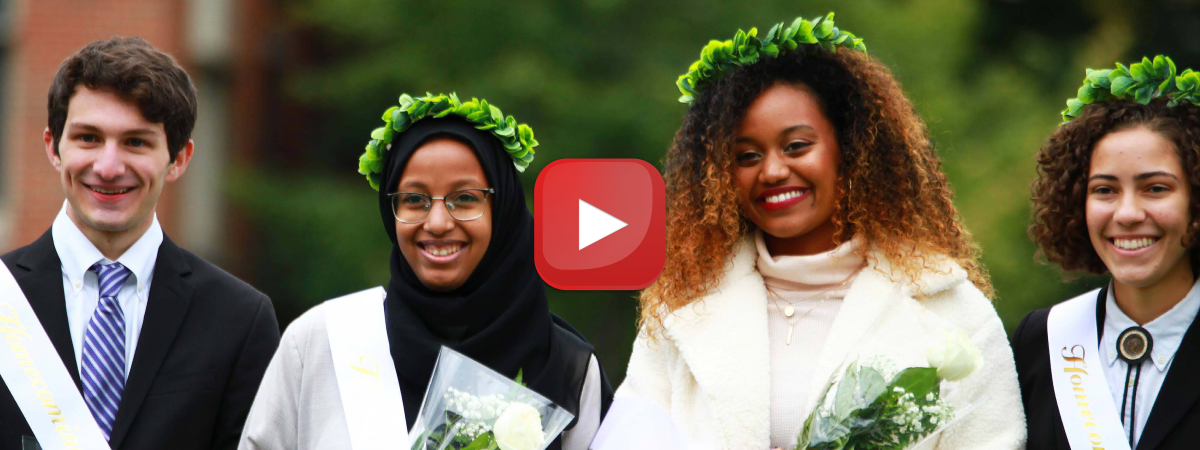 A diverse group of four CWRU students from the 2021 Homecoming Court stand smiling on the football field, holding flowers and wearing sashes, some of them wearing crowns made of greenery.