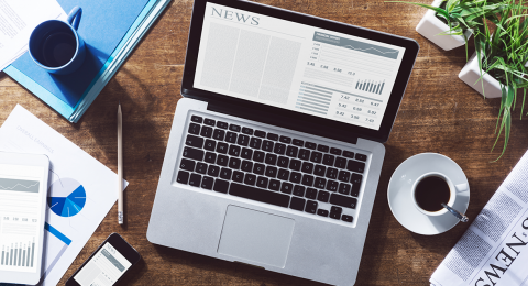 Photo of an open laptop with the word "news" on it, coffee cup, papers, newspaper and phone on a desk