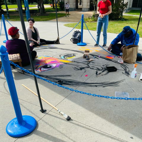 A bright blue chain surrounds Anna Arnold who is sitting on the ground with students on the edge of her outlined chalk portrait