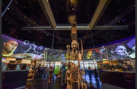 Inside the African American Museum of History in DC, an African sculpture stands in the center of a room with large screens showing famous Black musicians