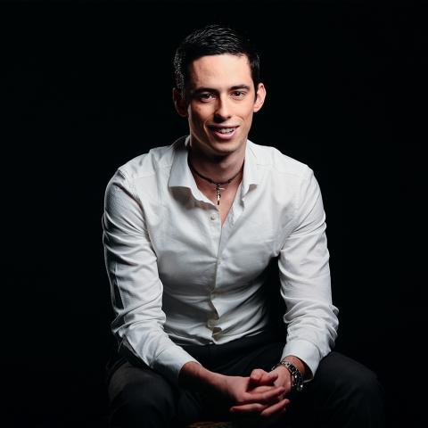 Felipe Gomez del Campo smiles, sitting down wearing a white shirt and black slacks with his hands clasped and arms resting on his knees