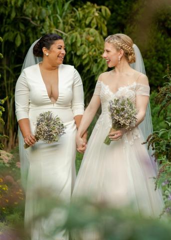 Rachel and Aquene Payne walk outside holding hands, wearing their wedding dresses, veils and carrying bouquets
