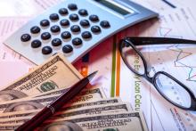 Photo of a stack of money, calculator, glasses all laying on top of budgeting documents