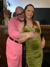 J Davenport poses in a green dress with her partner of 11 years, Trae Ruscin, in a bright pink jumpsuit embracing her.