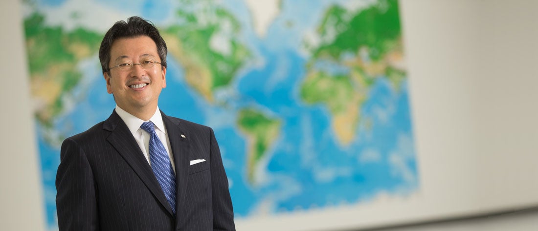 Hiroyuki Fujita wears glasses and a black suit and blue tie, standing in front of a world map