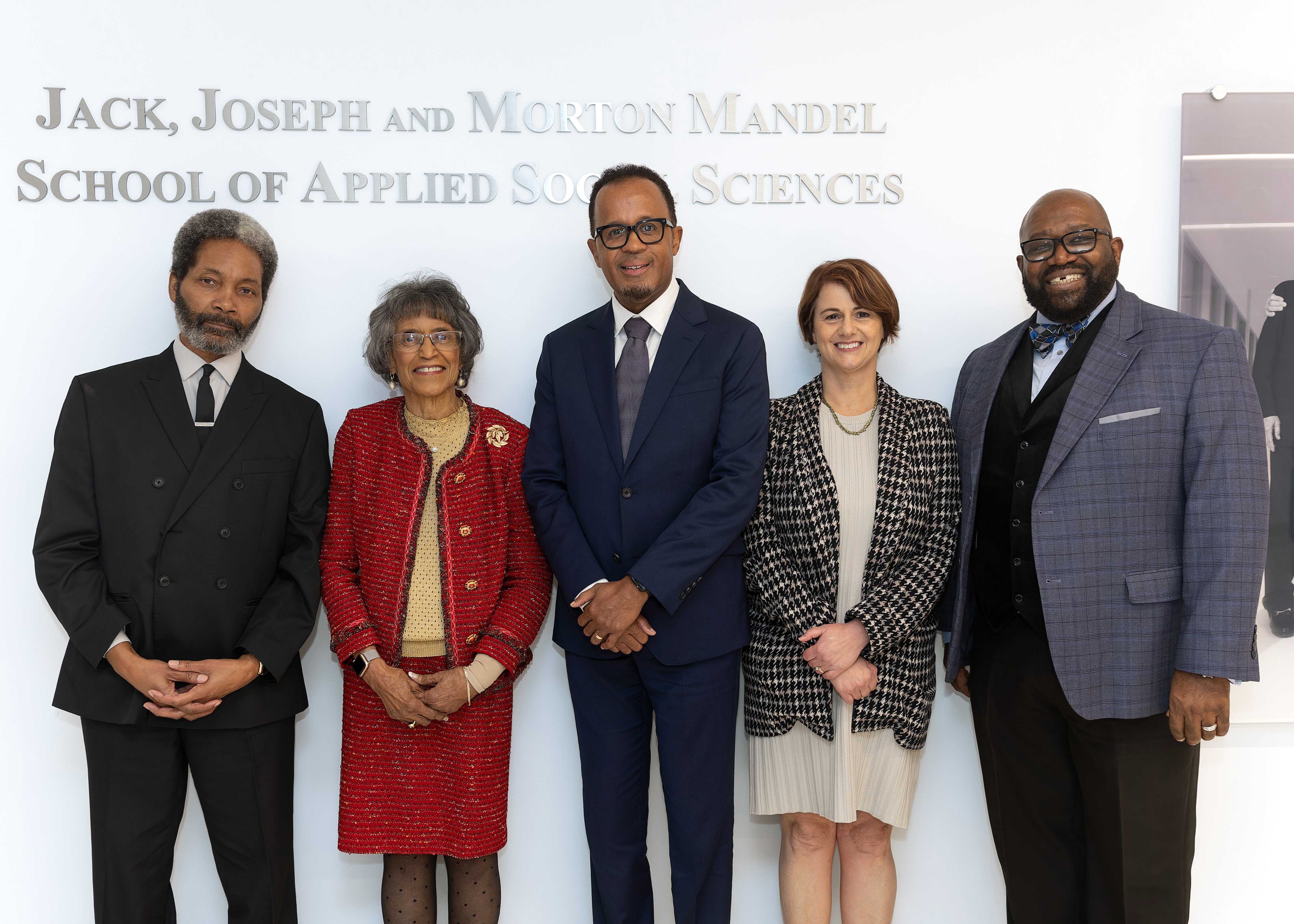 A group of five people dressed formally, in front of a wall that reads Jack, Joseph and Morton Mandel School of Applied Social Sciences