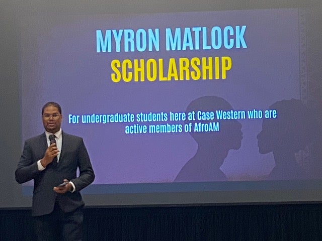 Myron Matlock presents about eponymous scholarship with presentation slide behind him, wearing formal attire