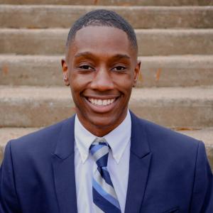 George Bukenya smiles at camera while wearing formal attire in front of outdoor concrete steps