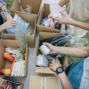 Foodbank volunteers sort grocery items into cardboard boxes at a table outdoors