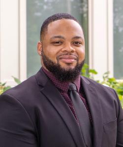 Murray wears a black suit jacket, maroon shirt and black tie, smiling at the camera
