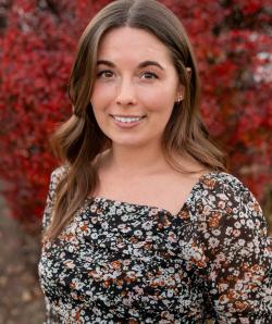 Brittany Hobel smiles at the camera in front of a tree with red leaves and wearing a floral blouse.