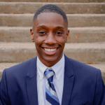 George Bukenya smiles at camera while wearing formal attire in front of outdoor concrete steps