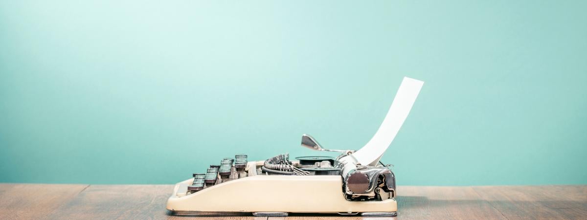 A small white typewriter sits on a table against a teal background
