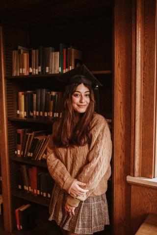 Sofia Castillo stands in front of a bookshelf while wearing a mortarboard cap, a sweater and a skirt