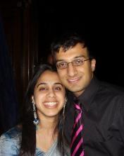A young Bhavani and Manu stand side-by-side, smiling brightly at the camera, Manu wearing a bright pink and black striped tie and Bhavani wearing long blue earrings.