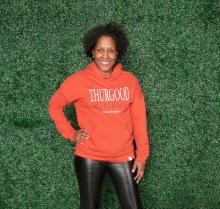 Michelle Felder wears a bright orange hoodie that reads "Thurgood Marshall," standing in front of a wall of greenery