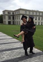 Man carries woman in an outdoor setting, both wearing graduation caps and gowns