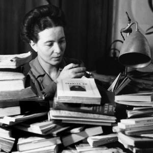 Black and white photo of Simone de Beauvoir at a desk surrounded by books