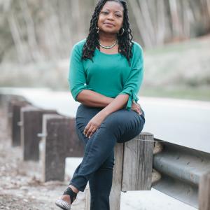 Image of an African American woman with long braided hair wearing a green sweater sitting on a guardrail
