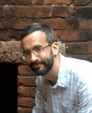 Color photo of white male with beard, mustache glasses in front of brick wall