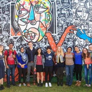 Color photo of group of diverse college students in front of a colorful outdoor mural
