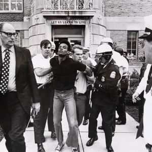 black and white image of a young African American man in handcuffs being moved by a crowd of white men including police officers 