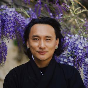 Head shot of Bhutanese man wearing black robe and blue earrings standing outside in front of flowering purple trees