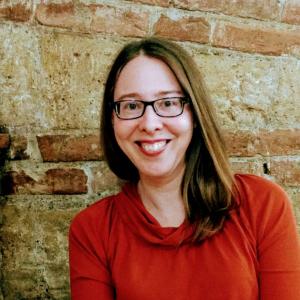 Image of smiling white woman with long brown hair, glasses, and a red sweater standing outside in front of a brick wall
