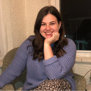 Smiling woman with long brown hair sitting in a chair resting her hand on her chin,